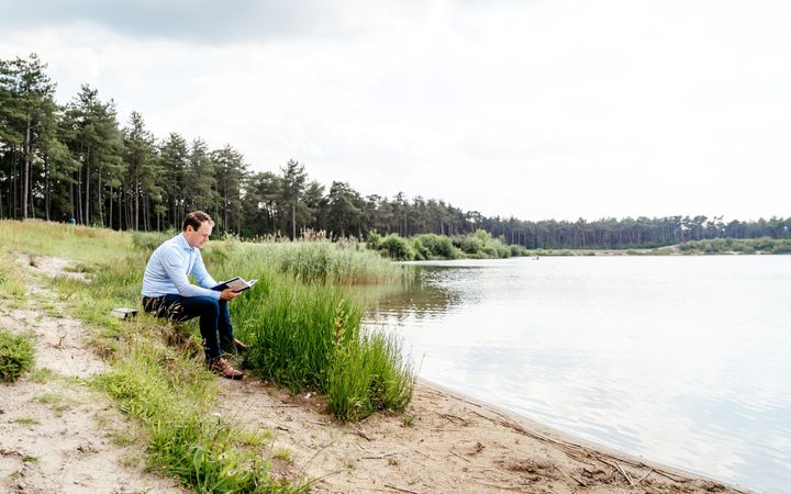 Jonge man met blauwe broek en lichtblauw overhemd, lezend aan een meer bij een bos.