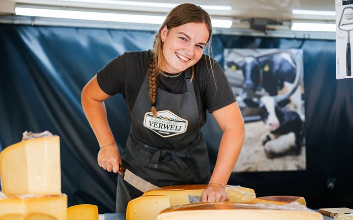 Jonge vrouw met zwart schort en zwart shirt snijdt met een groot kaasmes een stuk kaas af. Ze kijkt met een lach in de lens en heeft een lange vlecht in haar donkerblonde haar. 