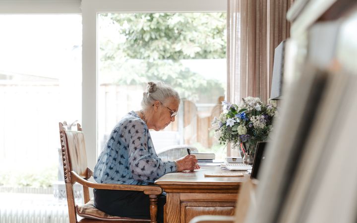 Oudere vrouw met grijs haar achter een bureau, terwijl ze iets opschrijft.