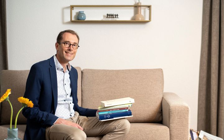 Man met lichte broek en blauw jasje, zittend op een bank met op zijn linkerbeen een stapel boeken.
