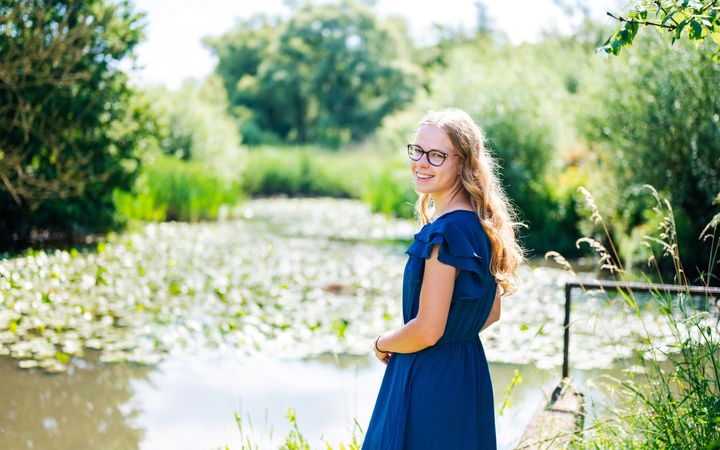 Jonge vrouw met blauwe jurk, lang, blond haar en een bril met donker montuur op.  Op de achtergrond een vijver met waterlelies. 