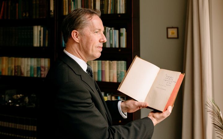 Man in donker pak voor een boekenkast met een boek in de hand.