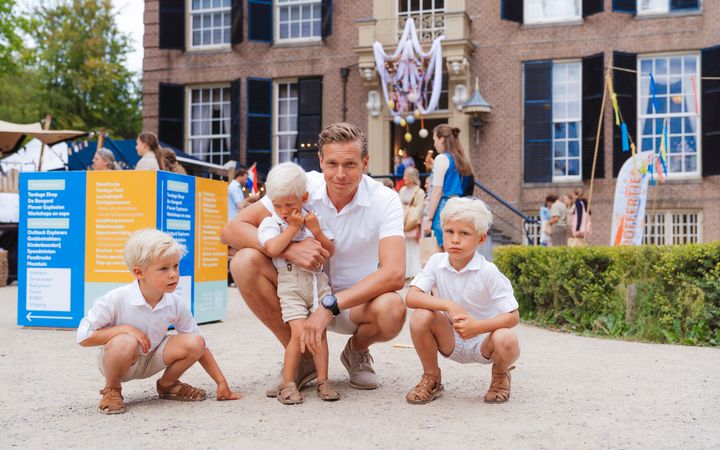 Vader met drie zoontjes in lichte, zomerse kleding. Op de achtergrond stands van exposanten, in een parkachtige omgeving. Rechts kleurige vlaggetjes.
