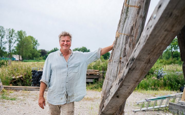 Man in kluskleding bij dikke houten balken, in een groene omgeving.
