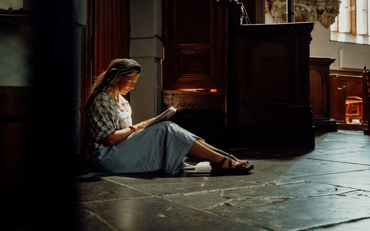 Vrouw met blond haar en een blauwe rok, lezend in een boek op de vloer van een oude kerk.