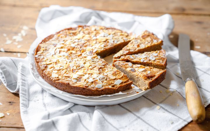 Wit bord op een wit-grijs geruite theedoek. Op het bord een platte, ronde en goudbruin gebakken koek met amandelschaafsel erop. 