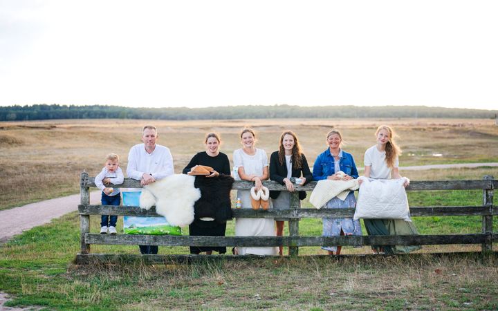 Houten hek met daarachter een kind, een man en een aantal vrouwen met wollen producten. 
