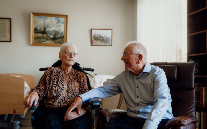 Oude vrouw in een rolstoel naast een oude man in een stoel, met een hand op haar hand.