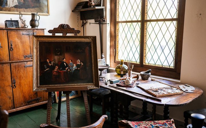 Antieke meubels, een ingelijst schilderij op een schildersezel en schildersbenodigdheden op een tafel voor een raam.