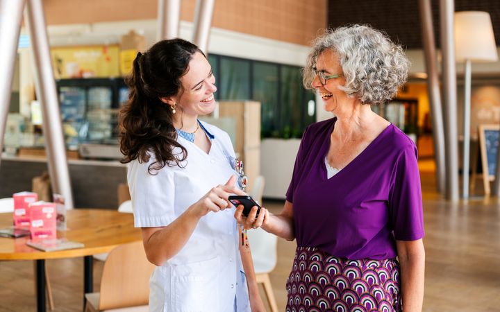Twee vrouwen staan in een ruimte. Een van hen heeft een mobiel in haar hand. 