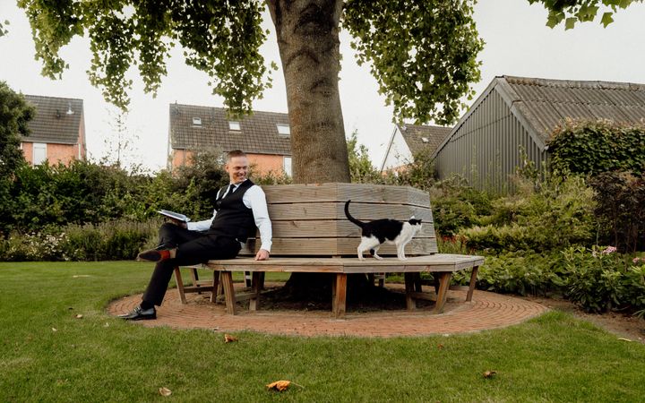 Man gekleed in zwarte pantalon, zwart gilet, zwarte das, wit overhemd en zwarte sokken en schoenen; zittend op een bankje onder een boom. Op het bankje loopt een zwart-witte kat. 