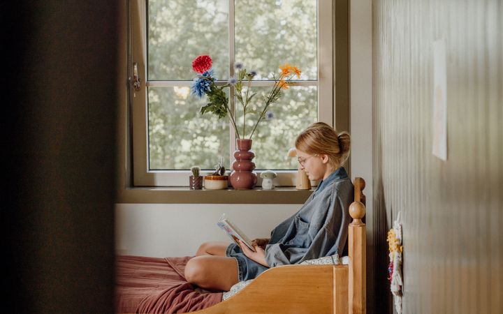 Een meisje zit op een bed, voor het raam, te lezen. De muren zijn oudgroen.
