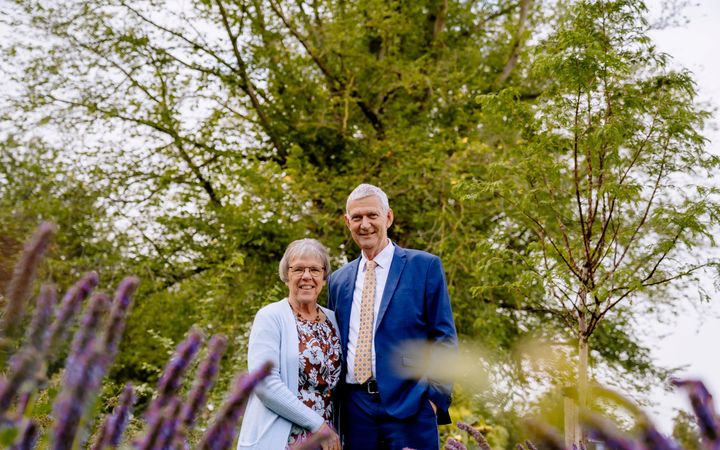 Vrouw met lichtblauw vest naast een man in een blauw kostuum in een tuin met bomen.