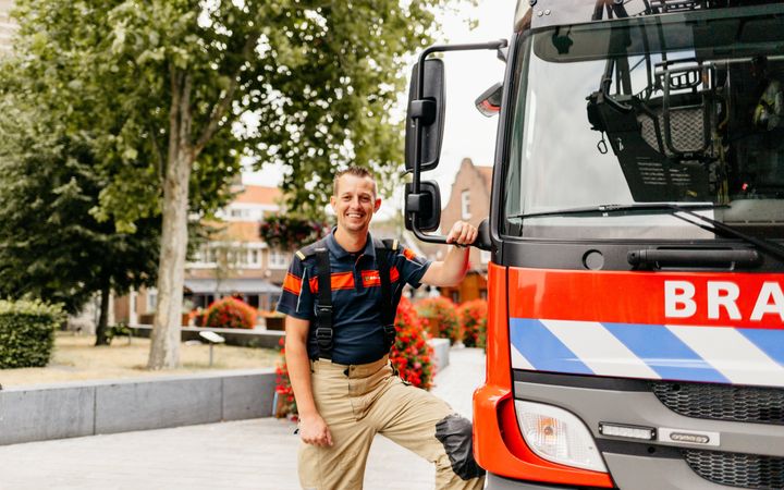 Brandweerman naast een brandweerwagen. Op de achtergrond bomen en huizen.